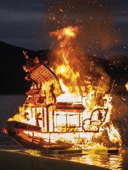 An image of a structure burning at night in a mountain lake setting, with two recording figures on either side
